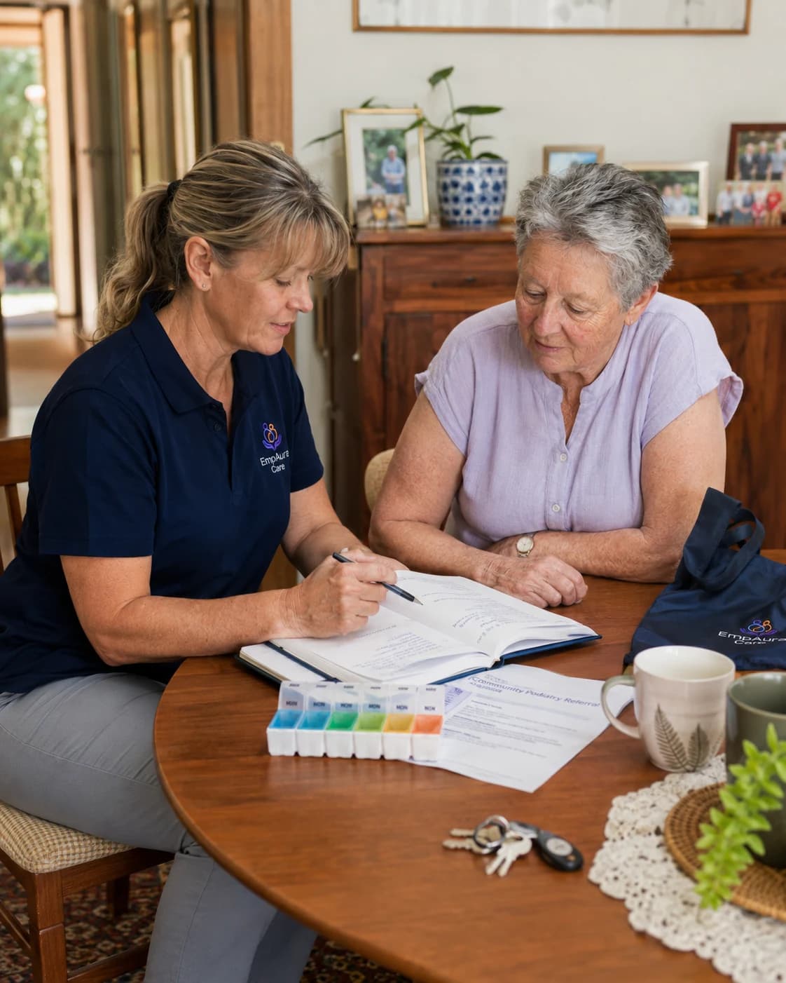 EmpAura Care community nurse reviewing a care plan with a participant at her kitchen table, with a weekly pill organiser visible